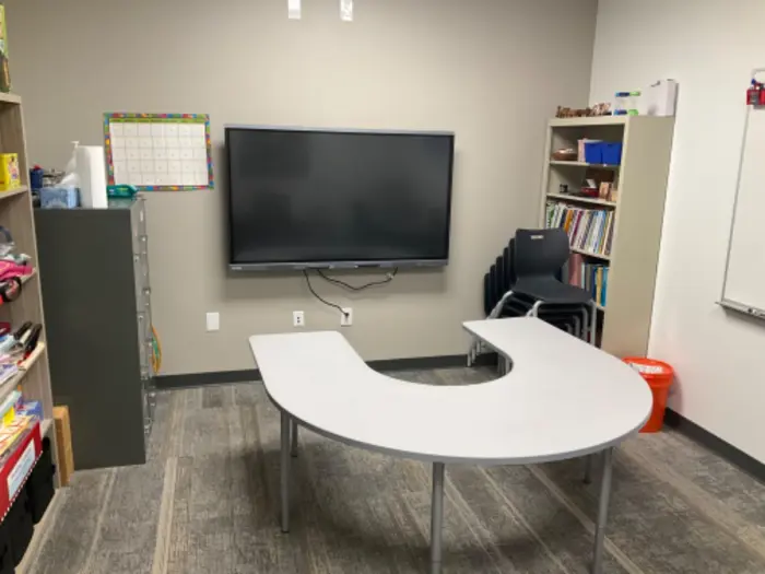 A small classroom with a U-shaped table and a large flat-screen TV on the wall is ideal for testing ideas. Shelves filled with books and supplies, a stack of chairs in one corner, and a calendar on the wall complement the gray carpet and light-colored walls.