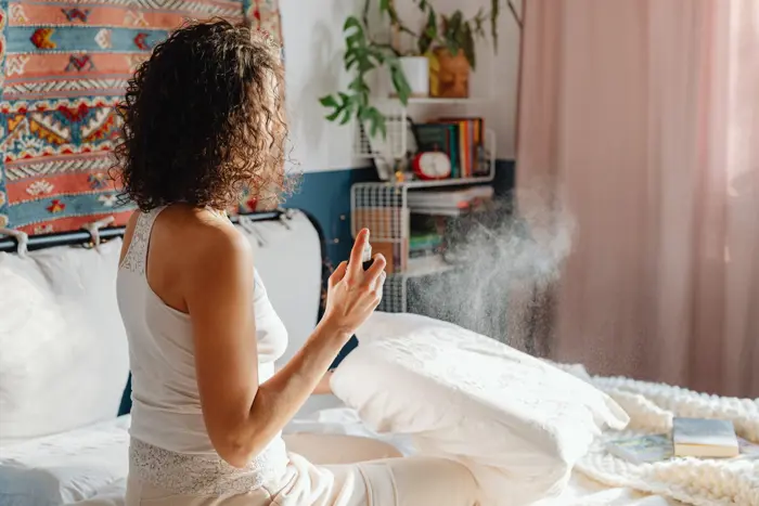 A person with curly hair sits on a bed, spraying a mist over a pillow, perhaps mindful of mold testing. The cozy room features a colorful tapestry on the wall, bookshelves with various items, and plants in the background. The person wears a white tank top and beige pants.