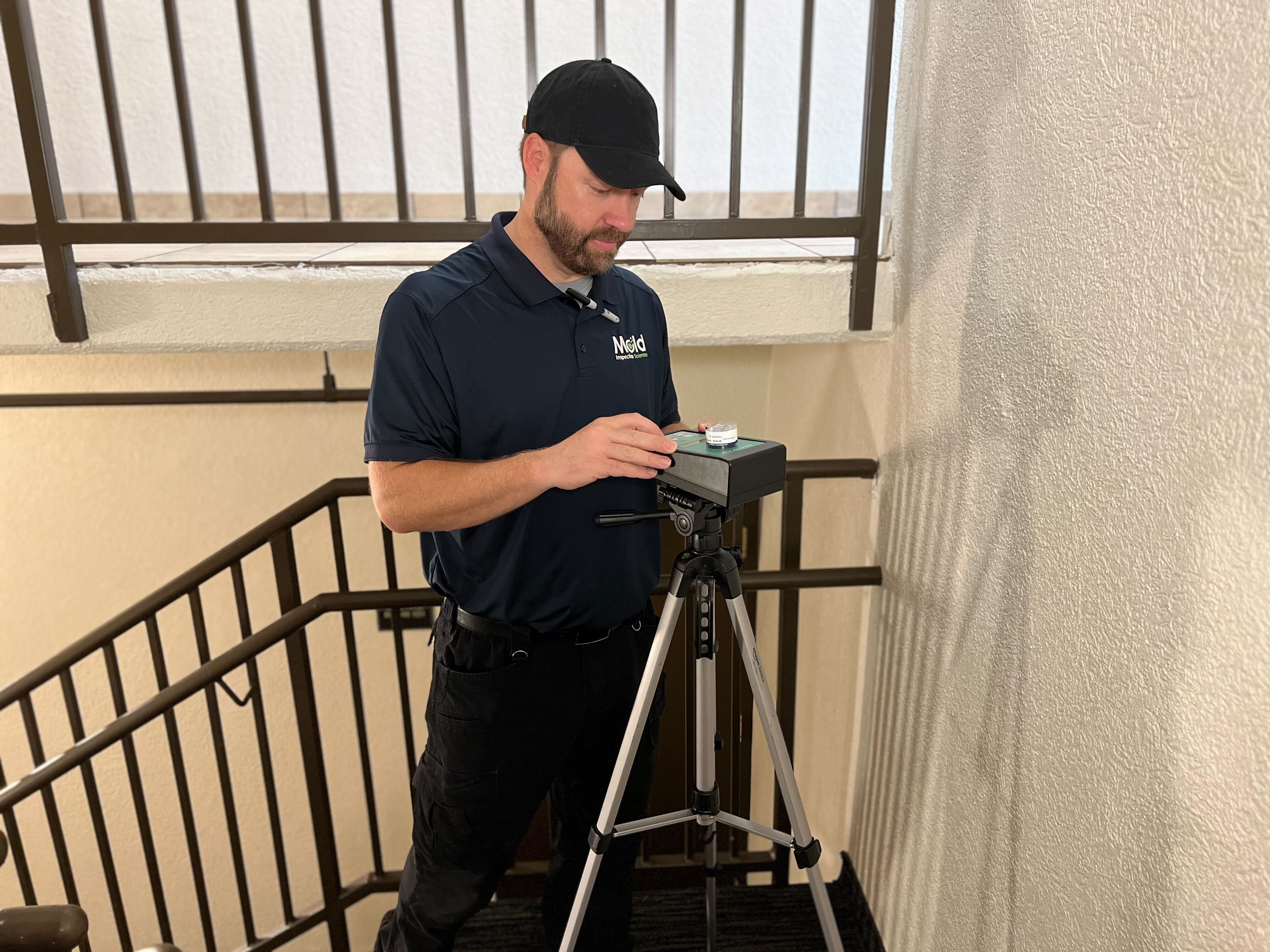 A man in a navy polo shirt and black cap adjusts equipment on a tripod for an inspection on a stair landing inside a building with beige walls and metal railings.
