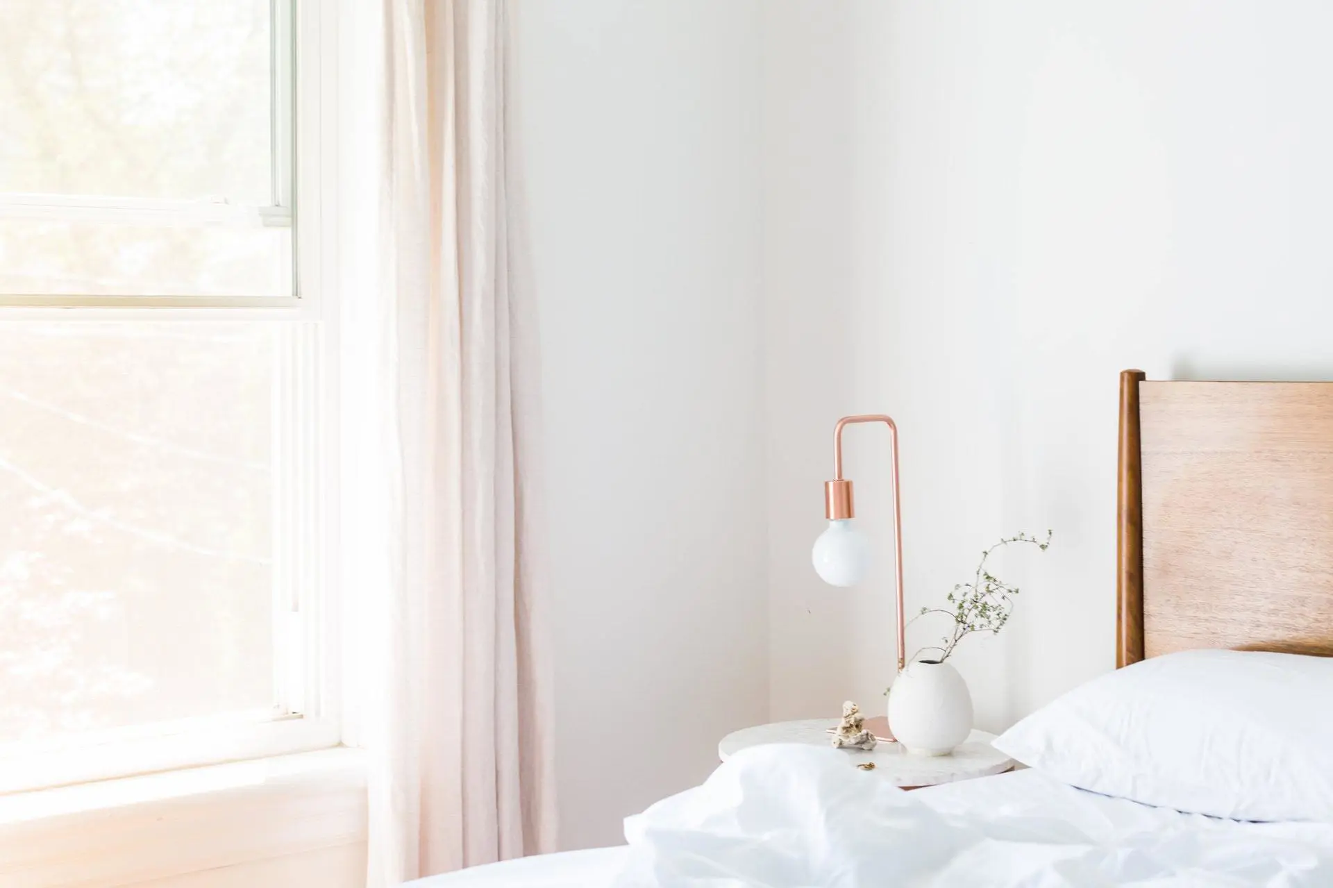 A bright and minimalistic bedroom with a wooden bed frame, white bedding, and a small round bedside table. The table holds a vase with small flowers and a stylish testing lamp. Soft natural light samples through sheer curtains at the window.