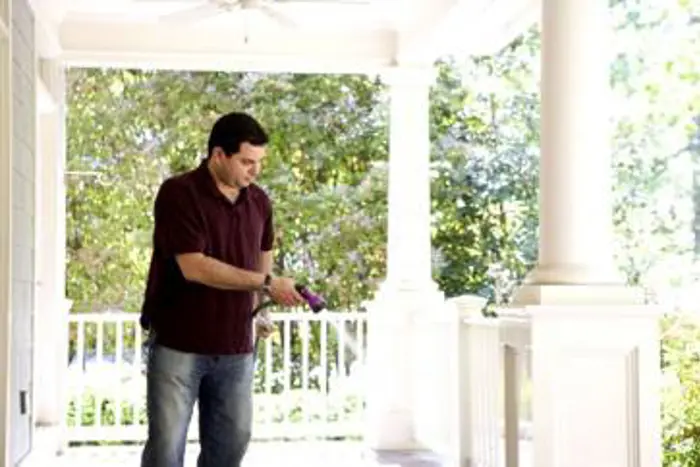 A man in a maroon shirt and jeans waters plants on a porch, inspecting each one with care. The setting is bright and sunny, with lush green trees visible in the background. The porch has white railings and pillars, creating a peaceful atmosphere perfect for his quiet testing of nature's beauty.