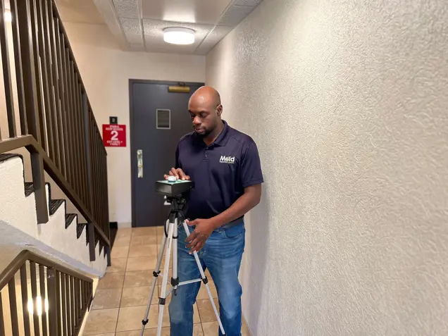 A person stands in a hallway, setting up a tripod-mounted camera for assessment. Clad in a dark polo shirt and jeans, they focus on ensuring everything is perfect. Stairs and a fire exit sign are visible in the background, setting the scene for this important task.
