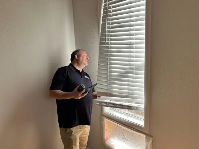 A person in a dark shirt and khaki pants smiles while inspecting the horizontal blinds on a window. They hold a tool in one hand as sunlight filters through, casting shadows on an object wrapped in plastic below.