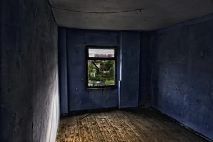 A dimly lit, empty room with dark blue walls and a wooden floor. A small window in the center offers a view of some greenery outside. The room has an abandoned and slightly eerie atmosphere, perfect for an investigation into its mysterious past.