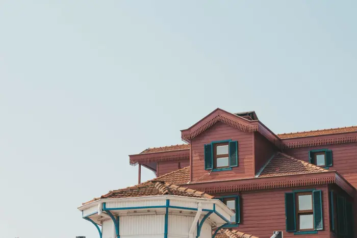 A reddish-brown, multi-story wooden house with green shutters and a triangular roof peak stands prominently. In the foreground, a smaller white structure with a round roof is partially visible, likely awaiting inspection. The sky is clear and light blue.
