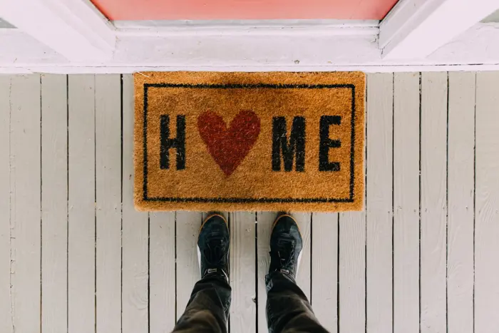 A welcome mat with the word HOME is shown, where the O is replaced by a red heart. A pair of feet in black shoes stands on the gray wooden porch, eagerly awaiting inspection before stepping into a warm embrace of comfort and security.