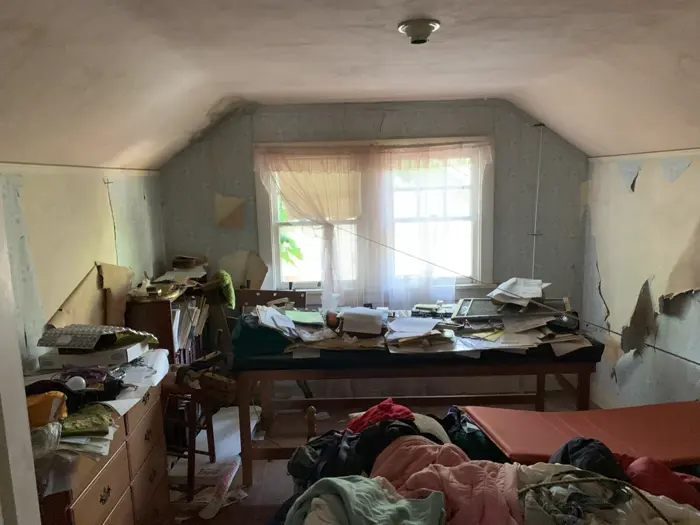 A cluttered, dimly lit room with peeling wallpaper suggests signs of wear. The bed is messy, and a desk is covered with disorganized papers and items, as if abandoned mid-investigation. A single window with sheer curtains allows some light in, highlighting the neglected state.