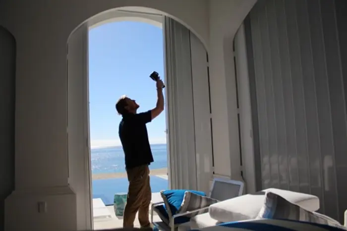 A person stands indoors near a large open doorway, silhouetted against a bright, sunny view of the ocean. They hold something up towards the light for assessment. Nearby are cushioned chairs and a table.