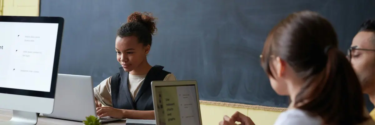 Three people are sitting at a table using laptops, with a large chalkboard in the background. One person faces the camera, while the others engage in sampling data. The room exudes a focused and collaborative atmosphere, perfect for this kind of investigation.