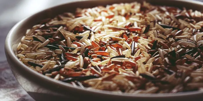 A close-up of a bowl filled with a mix of wild rice, where the long grains in shades of brown, red, and black invite closer inspection. Illuminated by soft natural light, their varied textures and colors reveal an intricate design worthy of deeper investigation.
