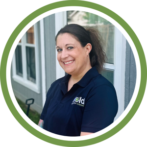A smiling woman with brown hair in a ponytail, wearing a navy polo shirt with a green and white logo, stands in front of a windowed building—framed by a green and white circular border—representing mold inspection services.