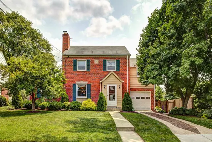 A two-story red brick house with white trim and green shutters is surrounded by a well-maintained lawn and trees. A concrete walkway, perfect for sampling the neighborhood charm, leads to the front door. There is an attached garage to the right with a short driveway.
