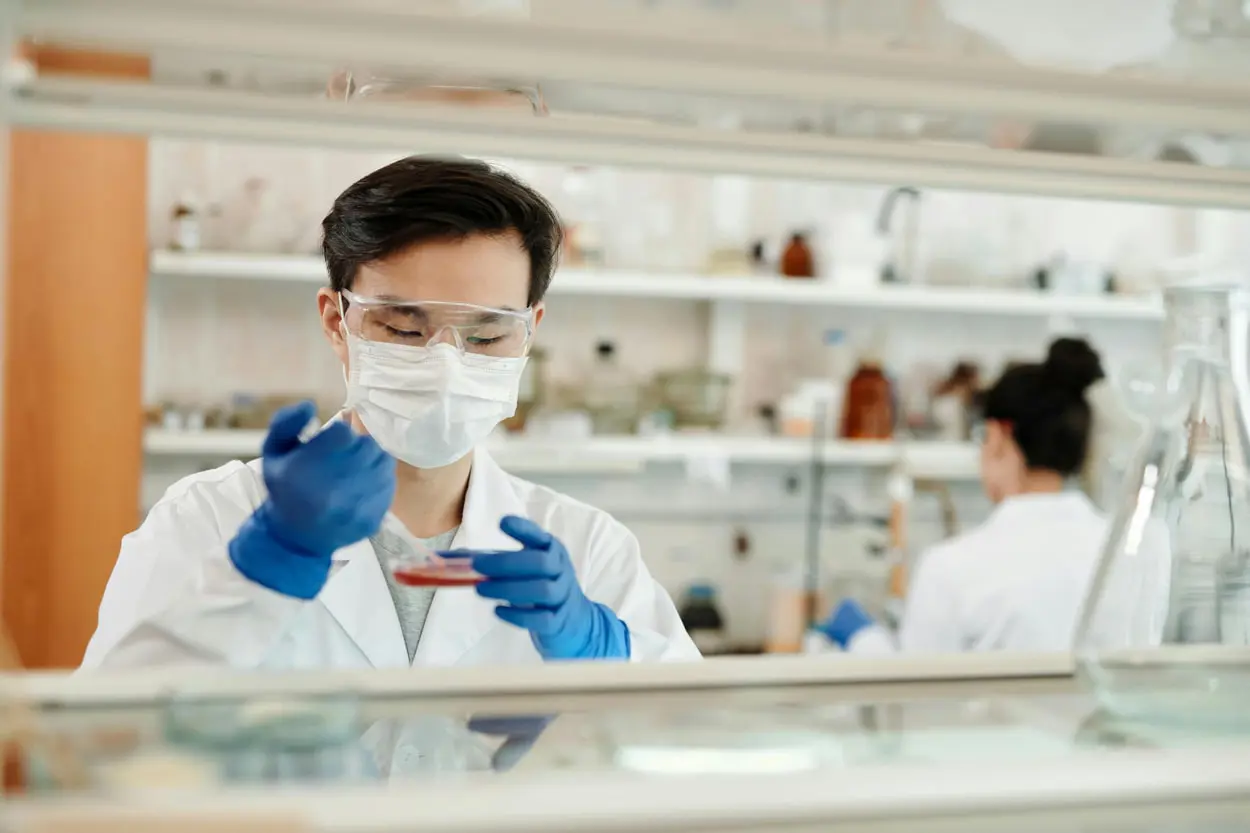 A scientist in a white coat, face mask, and safety goggles is focused on an investigation in the lab, carefully holding a petri dish with a pipette. Another researcher is visible in the background amidst shelves filled with lab equipment, possibly conducting mold sampling.