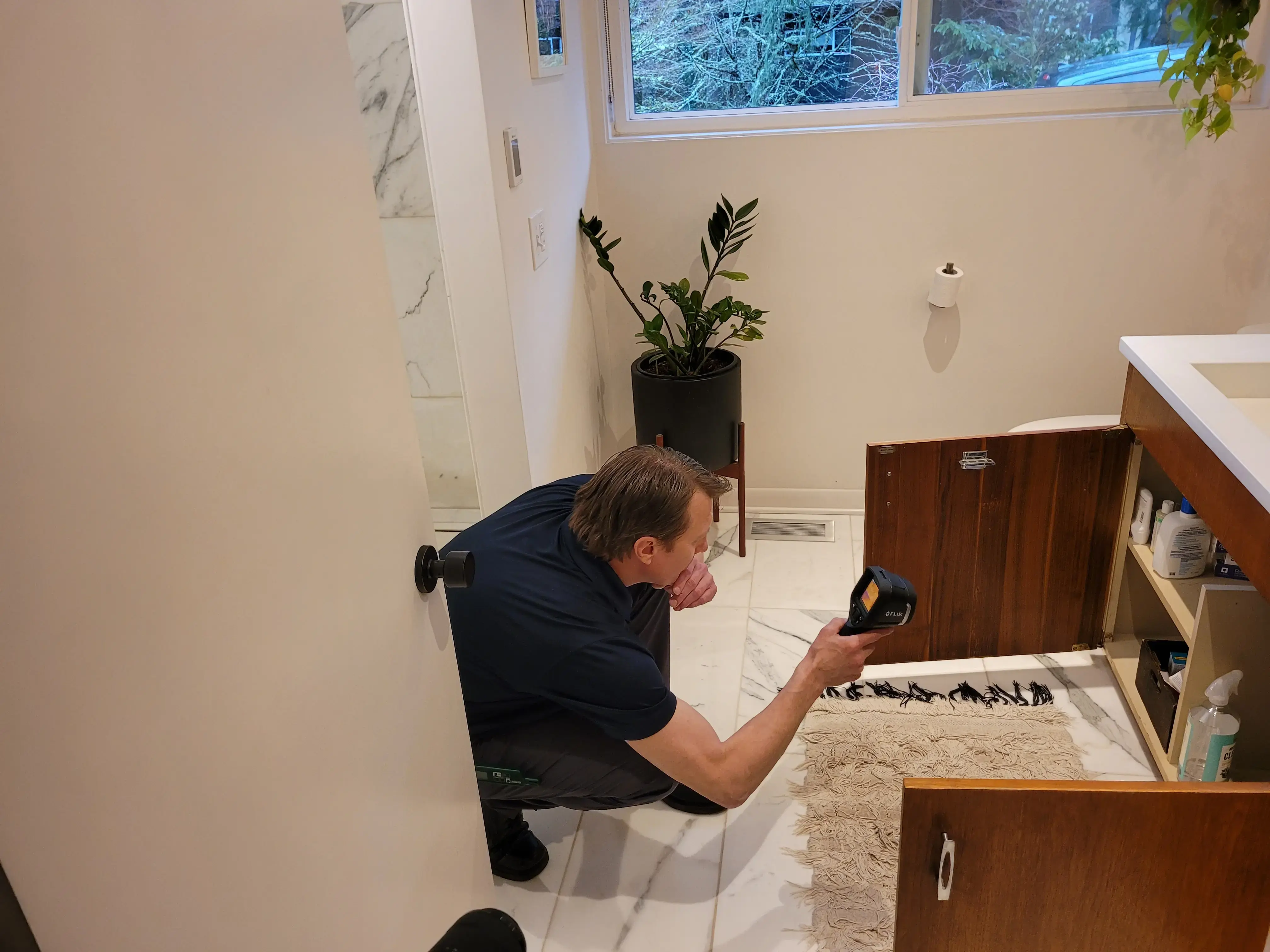A person kneels on the bathroom floor, using a flashlight to inspect for mold inside a cabinet beneath the sink. The bathroom features a white sink, a marble floor, a patterned rug, a potted plant, and a window.