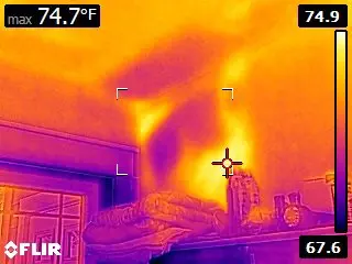 A thermal image displays a room with a bed and various items on a shelf. The temperature ranges from 67.6°F to 74.9°F, with hot spots on the wall suggesting areas for potential mold testing due to higher temperatures.