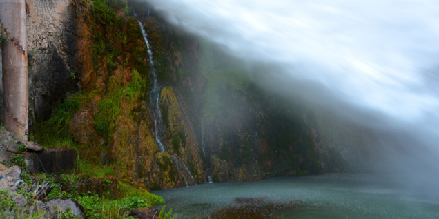 A small waterfall cascades down a rocky cliff, surrounded by lush green vegetation. Upon closer inspection, the water splashes into a calm pool below, creating a serene and misty atmosphere.