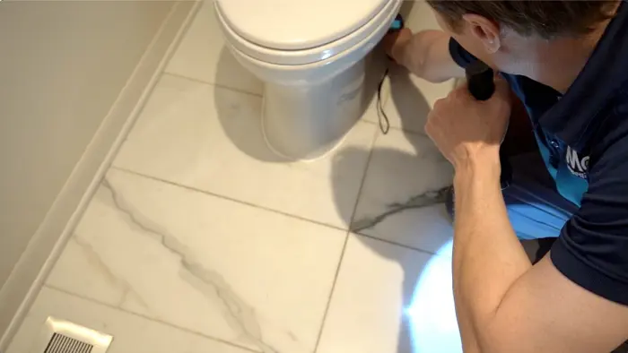 A person kneels on a marble-tiled floor, holding a flashlight and meticulously conducting an inspection of the toilet base. Clad in a blue uniform, they seem focused on assessing for any signs of mold or issues that require maintenance work.