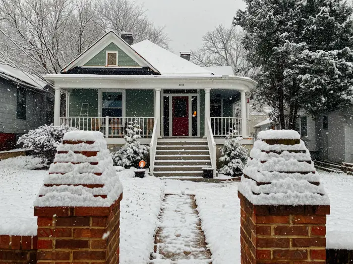 A cozy green house with a red door is blanketed in snow, evoking a peaceful and wintry scene. As snow covers the steps and yard, it's an ideal time for an indoor mold assessment. Two brick pillars frame the walkway while trees in the background are gently dusted with snow.