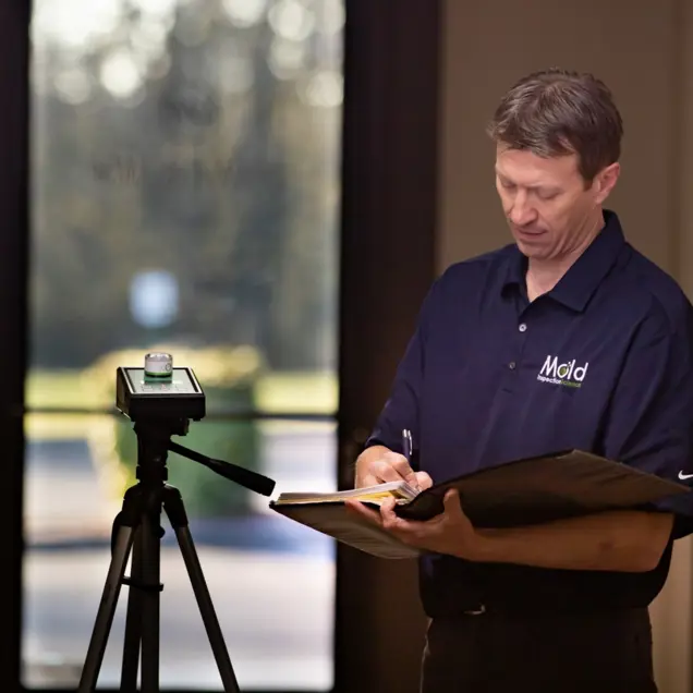 A man in a navy shirt stands indoors, conducting an assessment with a notepad and pen. He writes next to a tripod-mounted camera, perhaps documenting mold testing results. The blurred background features a doorway with outdoor light streaming in.