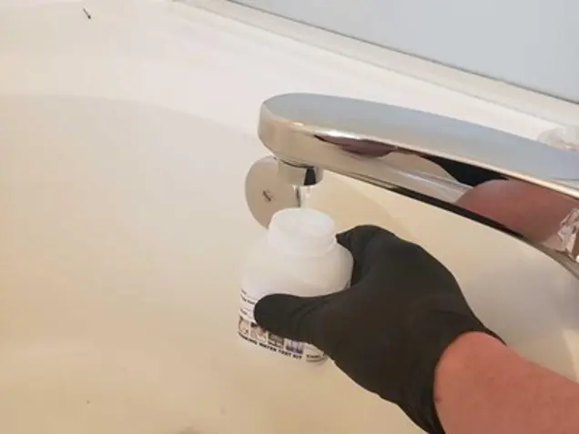 A person wearing a black glove holds an open plastic bottle under a running faucet, collecting water in a white bathroom sink, as part of an investigation.
