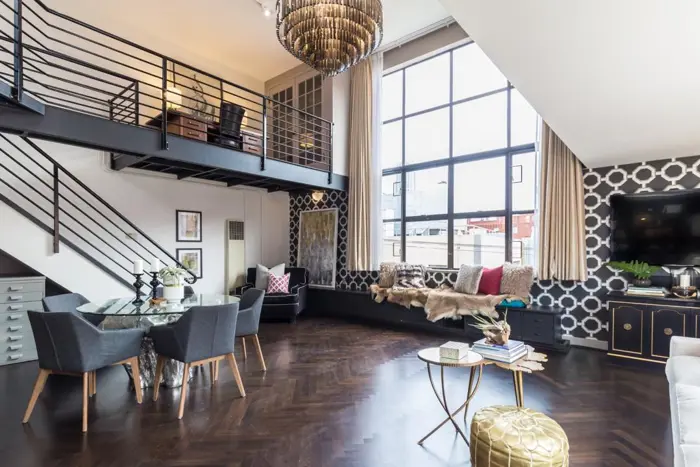 Modern loft with high ceilings and large windows. The space features a glass dining table, gray chairs, and a plush brown bench. A mezzanine with a desk offers an ideal spot for quick assessments, while the large chandelier illuminates wooden floors and geometric-patterned walls for a contemporary touch.