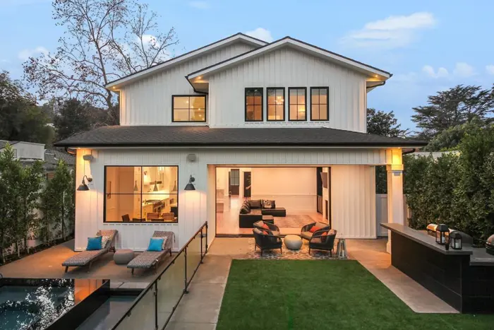 A modern two-story house with white siding and large windows. The backyard, inspected for safety, features a rectangular pool, lounge chairs, and a green lawn. The patio area is visible, offering a cozy outdoor seating arrangement with trees in the background.
