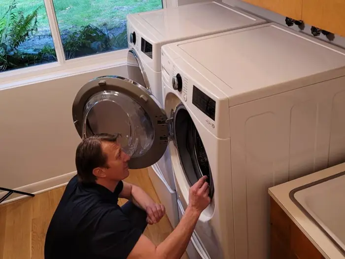 A person kneels in front of a white front-loading washing machine with the door open, conducting a thorough inspection of its interior. Another appliance stands nearby. The room features a large window revealing lush greenery, and part of a counter is visible on the right.