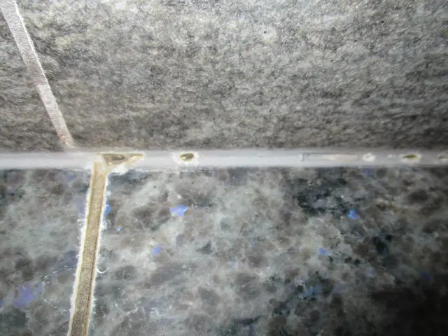Close-up of gray tiled surface with grout lines, ideal for a thorough mold investigation. The grout appears worn with small patches of discoloration. The tiles have a speckled texture, showcasing varying shades of gray with slight hints of blue.