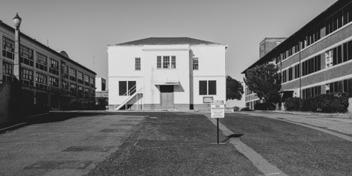 A black and white photo captures a small, white building with a staircase leading to the entrance, nestled between two larger buildings. The foreground features an asphalt area with a No Parking sign, resembling an urban investigation scene frozen in time.