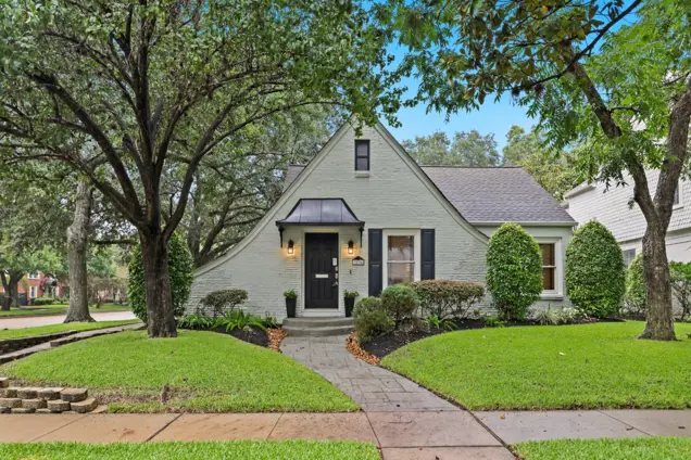 A charming cottage-style house with light gray brick, a steep peaked roof, and a black front door awaits inspection. Surrounded by lush green grass, manicured shrubs, and mature trees, it invites you in with a curved pathway leading to the entrance.