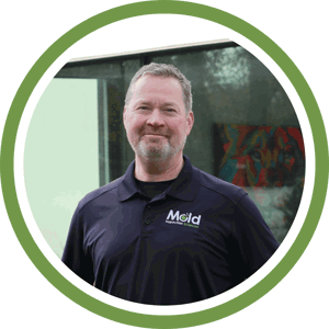A man with short hair and a beard is smiling at the camera. He is wearing a navy blue polo shirt with the word Mold embroidered on it, hinting at his expertise in mold assessment. The background features a glass window with reflections and greenery, framed by a green circle.