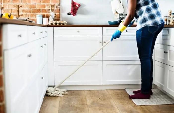 A person wearing a plaid shirt, jeans, and gloves is meticulously mopping a wooden kitchen floor, as if conducting an inspection. The kitchen features white cabinets, a brick wall, and various items on the counter. Nearby, a small white rug adds a touch of softness to the space.