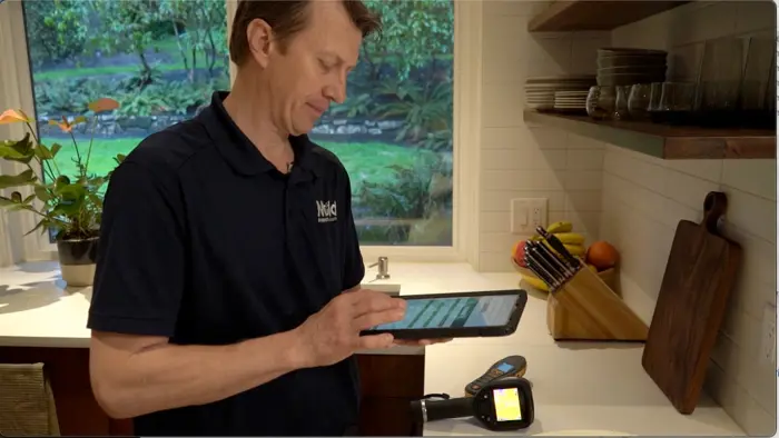 A person in a kitchen uses a tablet, wearing a dark shirt with a logo. On the counter, alongside various kitchen items, lies a digital thermometer for sampling ingredients. Through the window, lush greenery offers a serene backdrop to their culinary investigation.