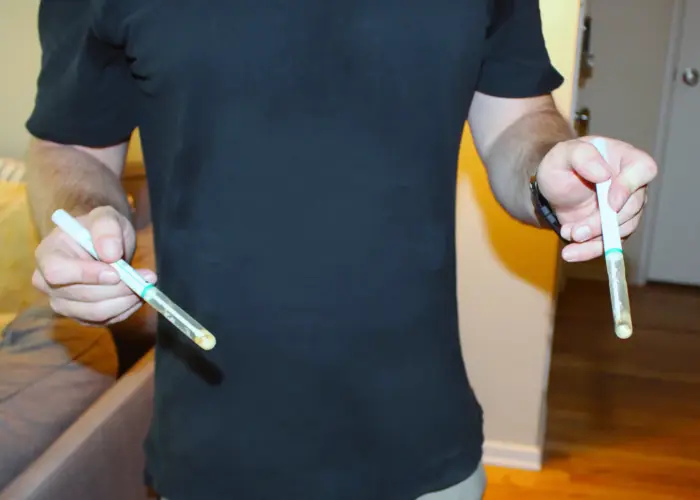 A person in a black shirt holds two test tubes containing a yellowish substance, one in each hand, as they conduct a testing session indoors beside a sofa and a doorway.