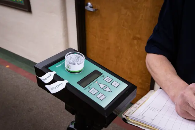 A person in a blue shirt holds a clipboard, conducting an assessment beside a green and black device labeled BIO-PAT with buttons and two small containers on top. The background reveals a wooden door and a beige wall, setting the scene for this precise investigation.