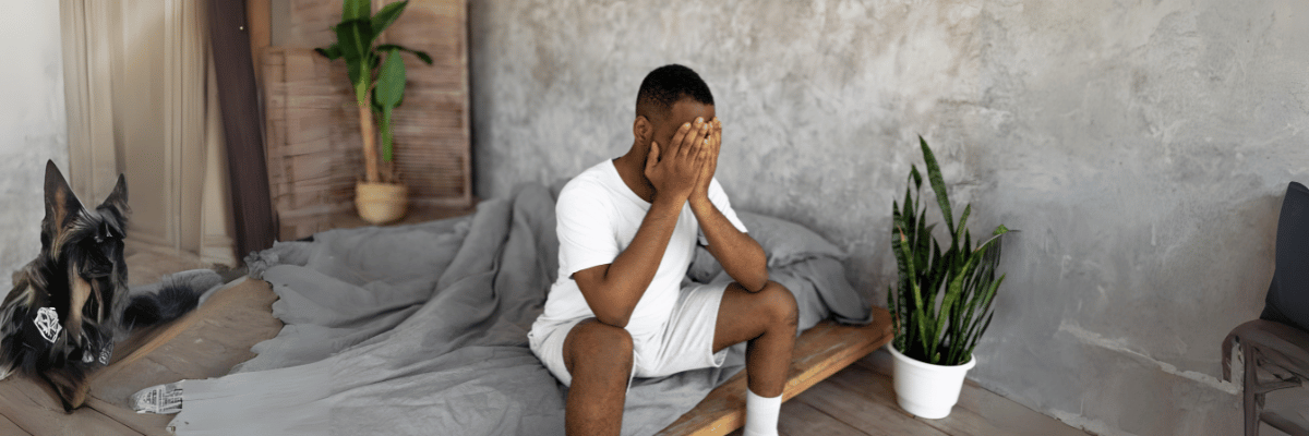 A person in a white outfit sits on a bed with their hands covering their face, appearing distressed, possibly about mold testing. A dog lies nearby, and a potted plant is on the floor beside a gray wall. The room has wooden flooring and a curtain in the background.