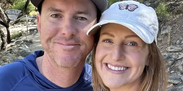 A smiling couple poses outdoors among trees. The man wears a blue shirt, sunglasses, and a cap; the woman, sporting a tattoo on her arm, wears a light tank top and a cap with a butterfly patch—ready for their next trail investigation.