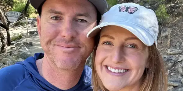 A smiling couple poses outdoors among trees. The man wears a blue shirt, sunglasses, and a cap; the woman, sporting a tattoo on her arm, wears a light tank top and a cap with a butterfly patch—ready for their next trail investigation.