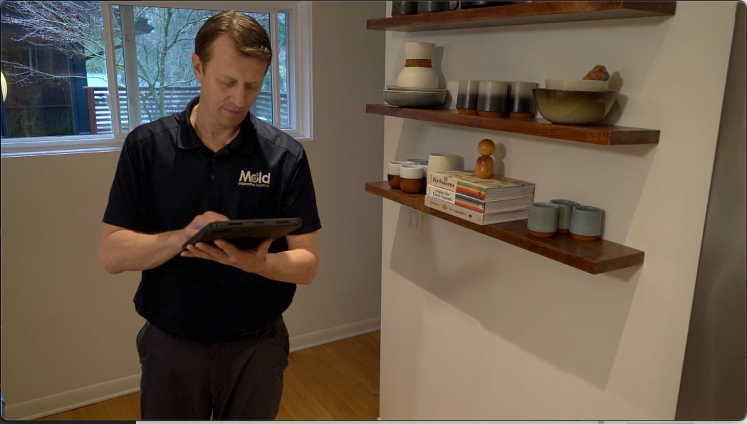 A person in a dark shirt holds a tablet, perhaps for inspection, while standing in a well-lit room with wooden floors. Behind them, wooden shelves on a white wall display various bowls, books, and decorative items. A window reveals trees outside.
