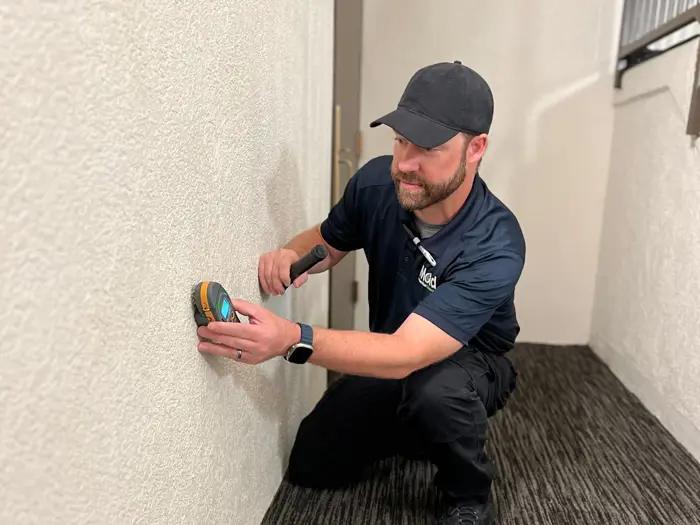 A person wearing a black cap and navy blue shirt crouches by a beige wall, conducting an inspection with a stud finder. They hold a flashlight and wear a smartwatch. The floor is carpeted in a striped pattern.