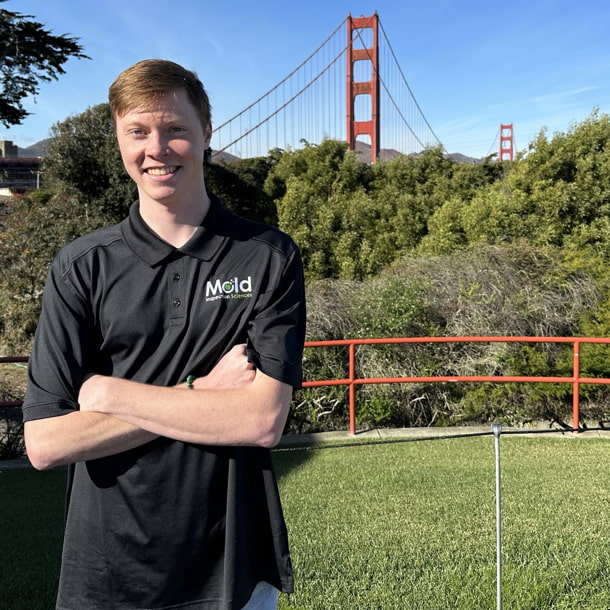 A person stands confidently with crossed arms, wearing a black polo shirt with a logo. They are in front of a scenic background featuring the Golden Gate Bridge and lush greenery, contemplating their next sampling project under the clear blue sky.