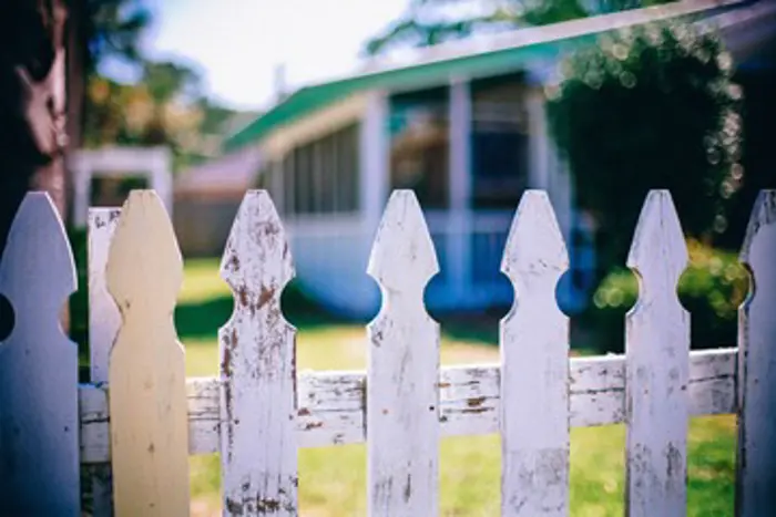 A close-up of a weathered white picket fence with peeling paint hints at years of exposure, inviting inspection. In the background, a blurred view of a house with a porch is surrounded by green grass and trees under a bright, sunny sky.