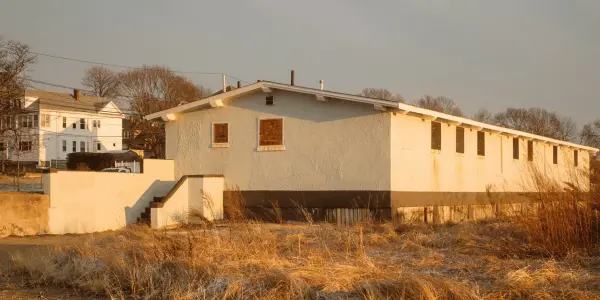 A long, single-story, white building with a flat roof sits in a field of dry grass, resembling a pristine canvas. Nearby are two multi-story houses. The scene is bathed in warm, golden light, perfect for an assessment of the area's beauty. Bare trees are visible in the background.