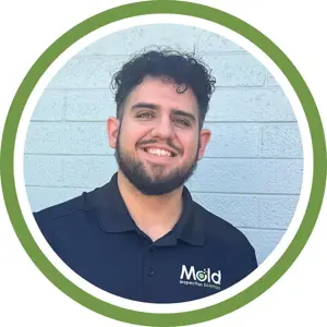 Smiling man with curly hair and beard wearing a black Mold Inspection Sciences shirt, standing in front of a light blue brick wall, framed by a green and white circular border—ready to handle your next inspection or assessment with professionalism.