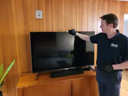 A person in a navy uniform and gloves is carefully sampling dust while cleaning a flat-screen TV on a wooden stand, surrounded by rich wooden paneling. Using a cloth, they inspect every corner of the screen, ensuring it's spotless as the TV remains turned off.