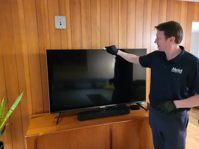 A person in a navy uniform and gloves is carefully sampling dust while cleaning a flat-screen TV on a wooden stand, surrounded by rich wooden paneling. Using a cloth, they inspect every corner of the screen, ensuring it's spotless as the TV remains turned off.