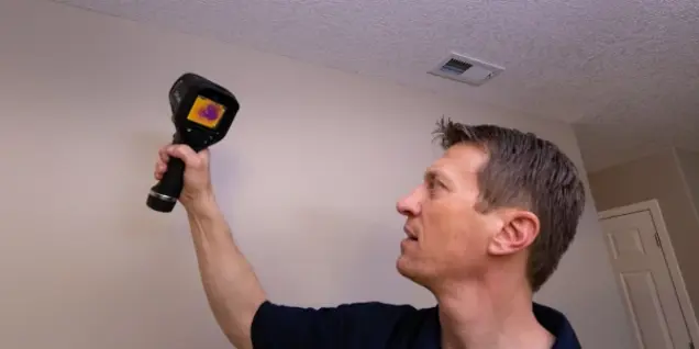 A person holds a thermal imaging camera up toward a ceiling vent, conducting an inspection of the wall and ceiling for temperature differences.