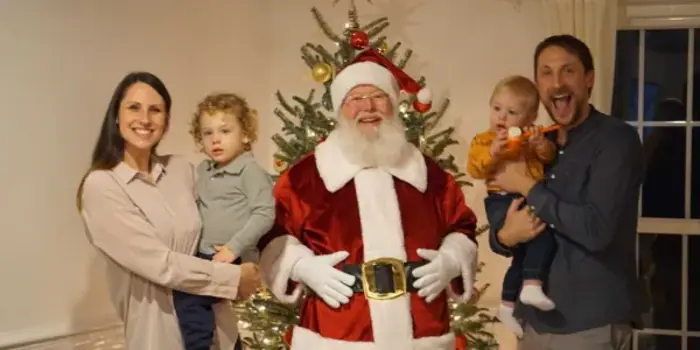A family with two children stands cheerfully with Santa Claus in front of a beautifully decorated Christmas tree. Their festive attire is impeccable, passing any holiday spirit inspection. The mother holds one child, while the father, on the right, holds the other. Smiles abound in this joyful scene.