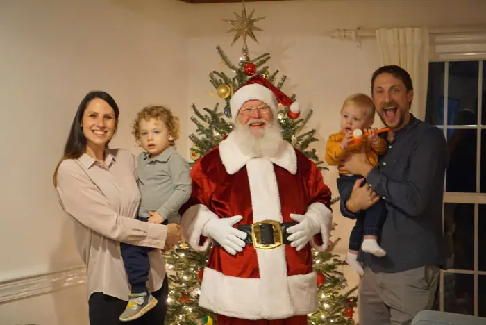 A family with two children stands cheerfully with Santa Claus in front of a beautifully decorated Christmas tree. Their festive attire is impeccable, passing any holiday spirit inspection. The mother holds one child, while the father, on the right, holds the other. Smiles abound in this joyful scene.