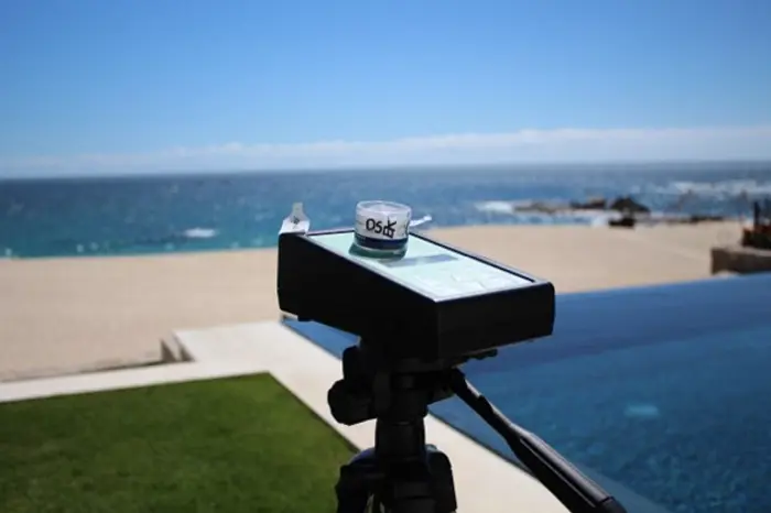 A camera on a tripod overlooks an infinity pool with a view of a sandy beach and the ocean, set for an investigative shot. A small lid labeled DSLR sits atop the camera. The sky is clear and blue, perfect for testing new photography techniques.